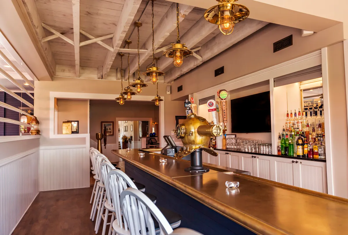 A stylish bar area featuring hanging pendant lights, a modern counter, and well-stocked shelves with drinks. The overall feel is relaxed and cheerful, suggesting a spot to unwind after a day at the beach or spa. The setup is conducive to friendly conversation or a quiet drink for two, reinforcing the sense of luxury and relaxation.