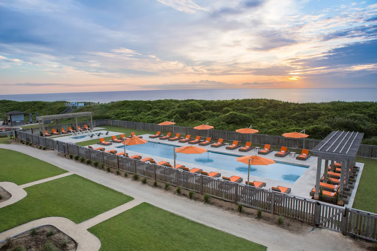 This image features a tranquil swimming pool area set against a sunset backdrop. Lounge chairs and orange umbrellas create a social and inviting atmosphere, ideal for groups to gather, relax, and enjoy each other's company. The warm, ambient light suggests evening swims and relaxed gatherings.