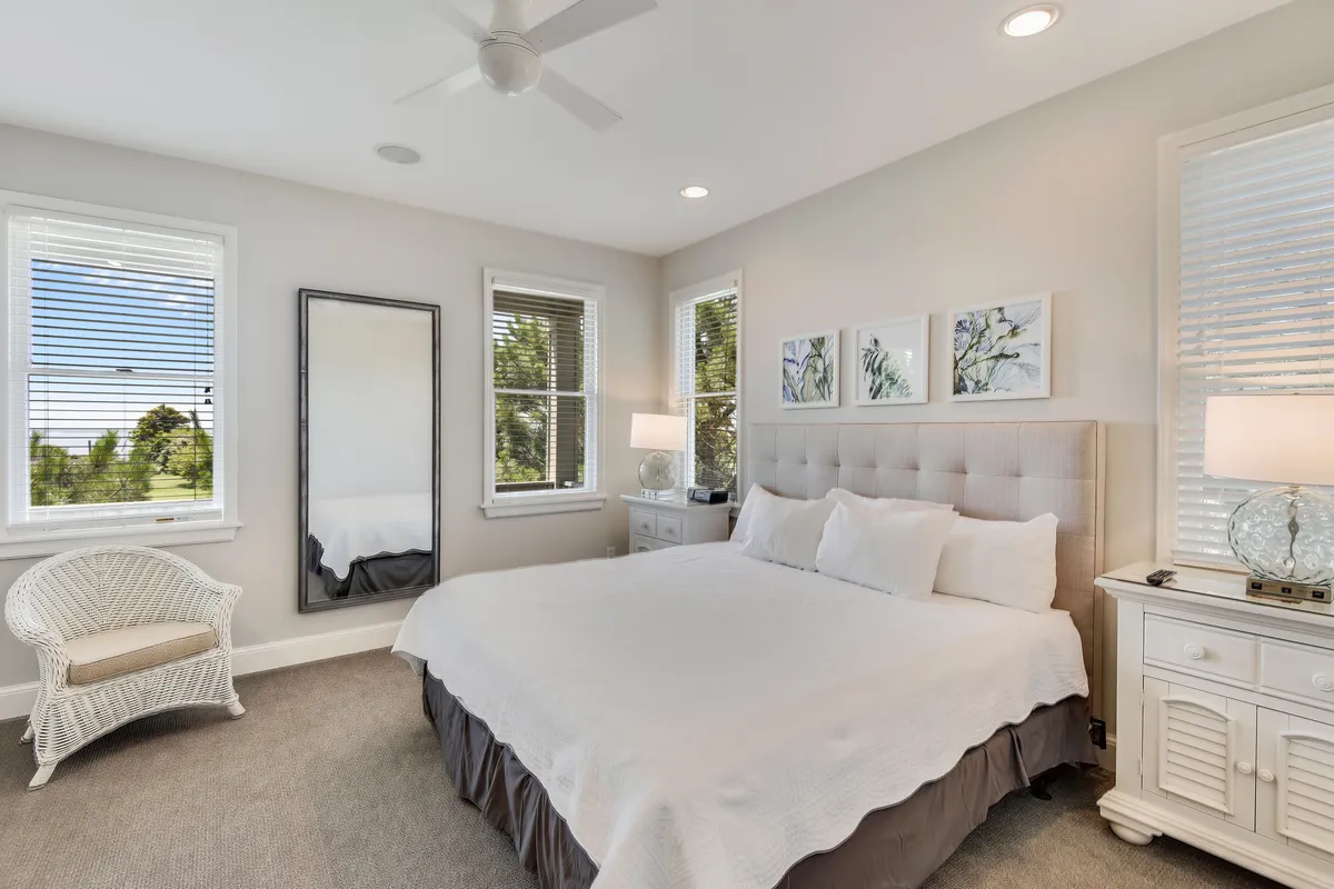 A serene bedroom featuring a king-sized bed dressed in luxurious linens, with decorative artwork above the headboard, a large mirror reflecting gentle light, a nightstand with a lamp for nighttime convenience, and a cozy wicker chair positioned near the window. The ambiance is calming and inviting, ideal for restful retreats.