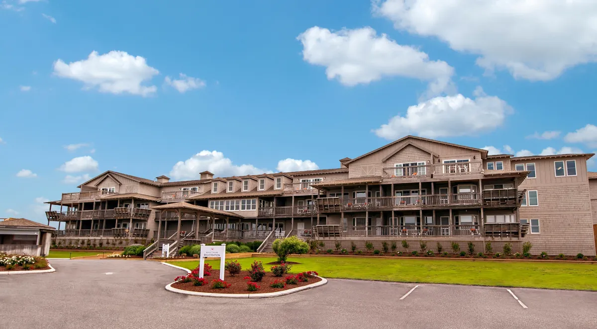 This image features the exterior of the Sanderling Resort, showcasing its inviting building facade, private balconies, and beautifully landscaped garden. The scene highlights people relaxing on the balconies, socializing in the communal garden, and enjoying the expansive outdoor environment. The lighting suggests either sunrise or sunset, underscoring the connection to coastal activities and natural beauty.