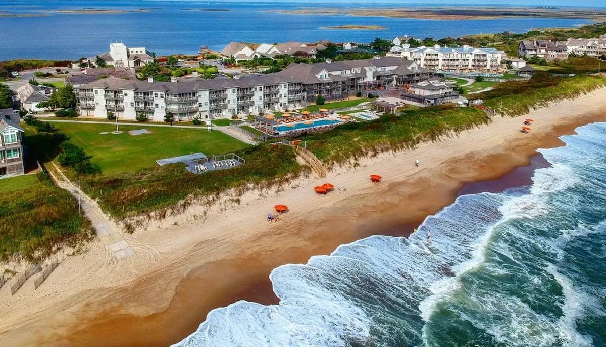A sweeping exterior view of the resort with direct beachfront access, a sparkling swimming pool, and hotel buildings overlooking the ocean. Umbrellas dot the sandy beach, inviting guests to lounge or stroll along the shore. The overall scene radiates a relaxed, high-end vacation vibe, especially with the sun low on the horizon.