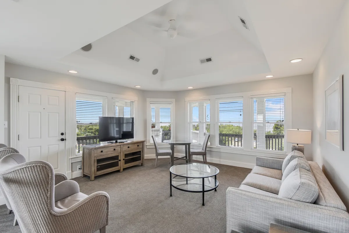 A spacious living area with plush sofas and armchairs arranged around a central coffee table. Large windows bathe the space in natural light, offering a soothing view of the outdoors. A television is positioned for entertainment, and the overall atmosphere is inviting and comfortable, ideal for relaxing or gathering with family and friends.