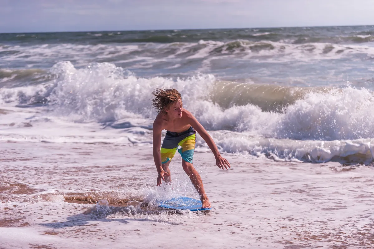 A joyful scene featuring a young boy surfing on a boogie board as waves crash around him beneath a vivid blue sky. The moment is full of energy, adventure, and the infectious fun of riding the surf.
