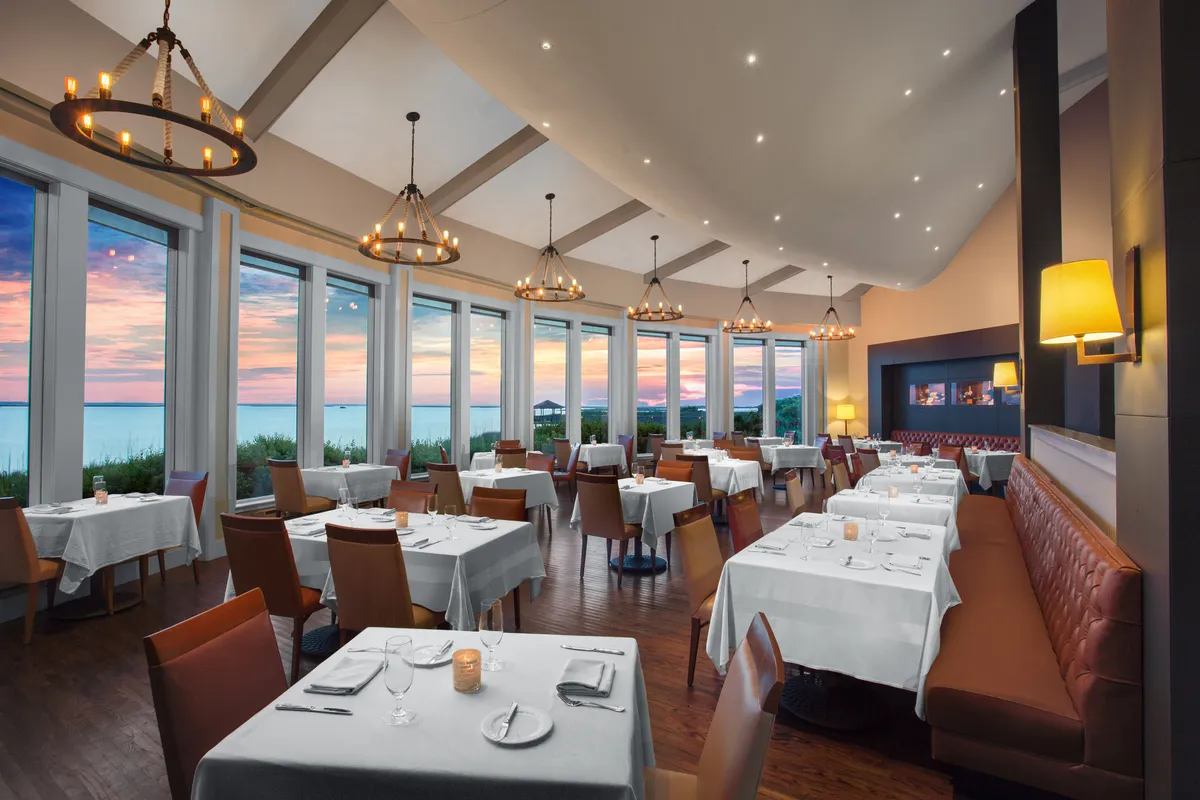 An elegant interior dining space with large windows framing an ocean view. Tables are set for fine dining under sophisticated chandeliers, and a cozy fireplace adds warmth to the atmosphere. The mood is refined yet relaxed, ideal for savoring gourmet meals or intimate gatherings.