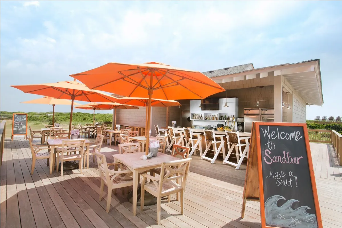 The scene captures an outdoor bar and dining area, complete with vibrant orange umbrellas, wooden tables and chairs, and a welcoming sign. The mood is cheerful, with guests dining, socializing, and enjoying views under open skies. The setting suggests a lively spot for winding down after a day of adventure, with easy access to fresh air and coastal ambiance.