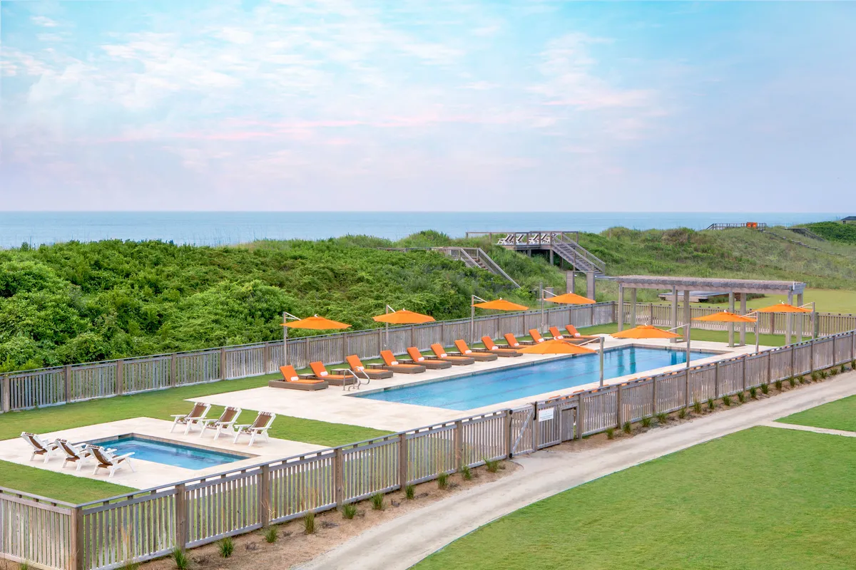 A tranquil exterior shot featuring a resort swimming pool lined with lounge chairs and umbrellas. The pool overlooks a stunning ocean view, and a winding pathway invites guests to explore the lush grounds. The mood is serene, with hints of social and wellness activities that guests can enjoy.
