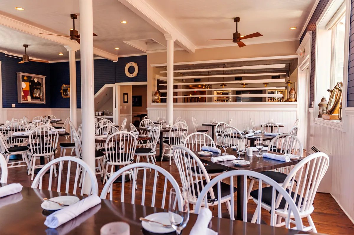 A bright, airy dining space with white wooden chairs and dark wooden tables arranged neatly throughout the room. The decorative wall art and open bar area create a welcoming atmosphere, while natural light pours in, suggesting proximity to the outdoors. Couples and small groups can easily envision themselves sharing a relaxed meal, with an ambiance that balances casual comfort and understated elegance.