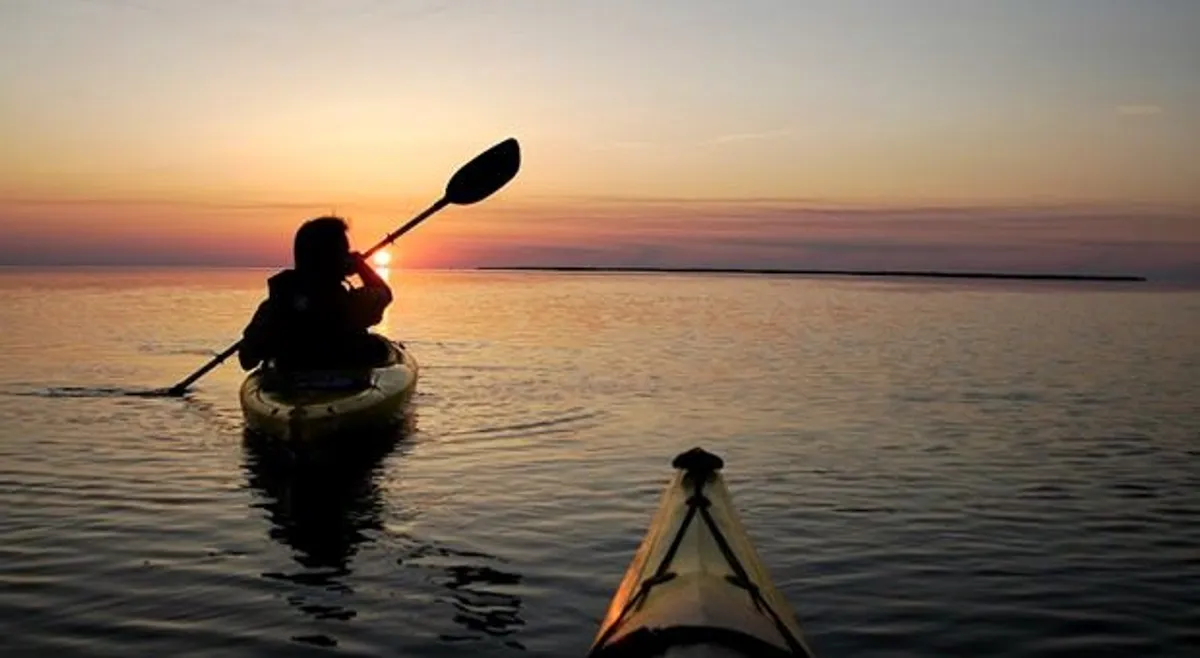 A kayaker glides across calm water at sunset, bathed in golden hues. The expansive view captures the tranquil beauty of the Currituck Sound, with the water reflecting the vibrant sky and creating a peaceful, immersive atmosphere.