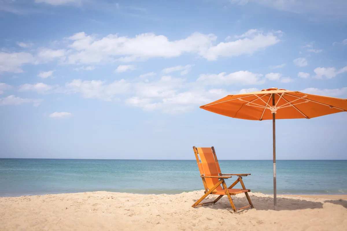 A tranquil stretch of sandy beach with an inviting orange beach chair and umbrella set up near the calm ocean. The scene is peaceful and bright, ideal for families ready to build sandcastles, read, or relax by the water under the shade of umbrellas.